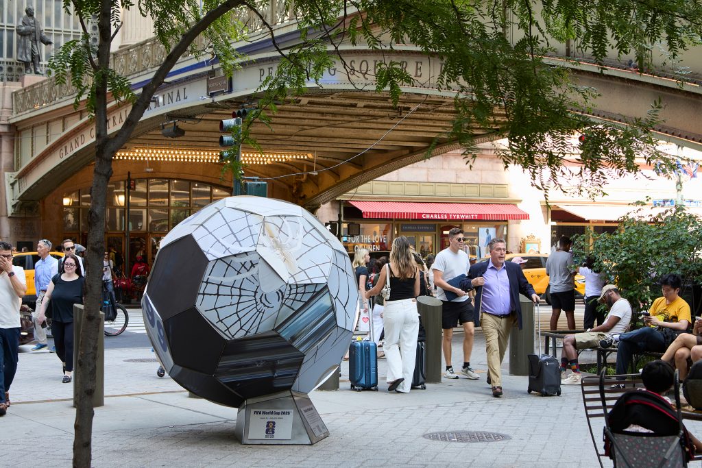 soccer ball sculpture with spider web pattern next to a bridge outside Grand Central Station with many people around it
