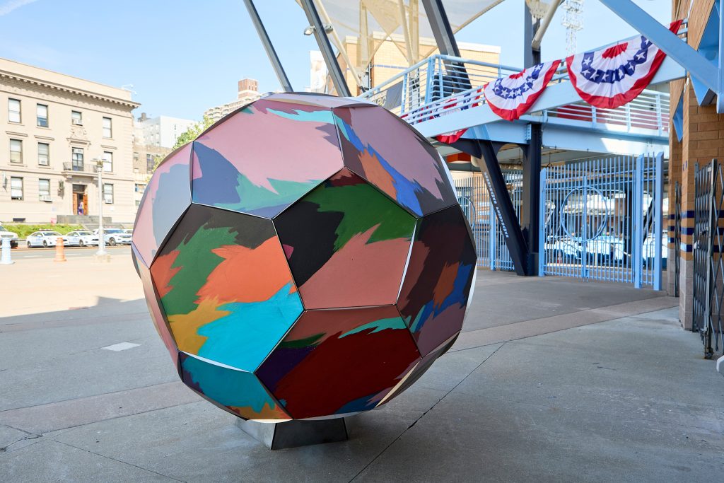large soccer ball sculpture with abstract color blocks painted on it in a plaza in front of a gate with bunting