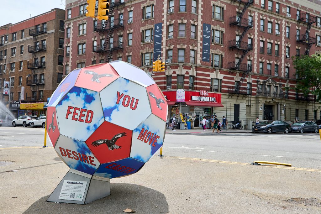 soccer ball sculpture by Cemile Sahin with some panels shoing birds and the others clouds and blue sky with words located on a plaza at a city street intersection in Washington Heights