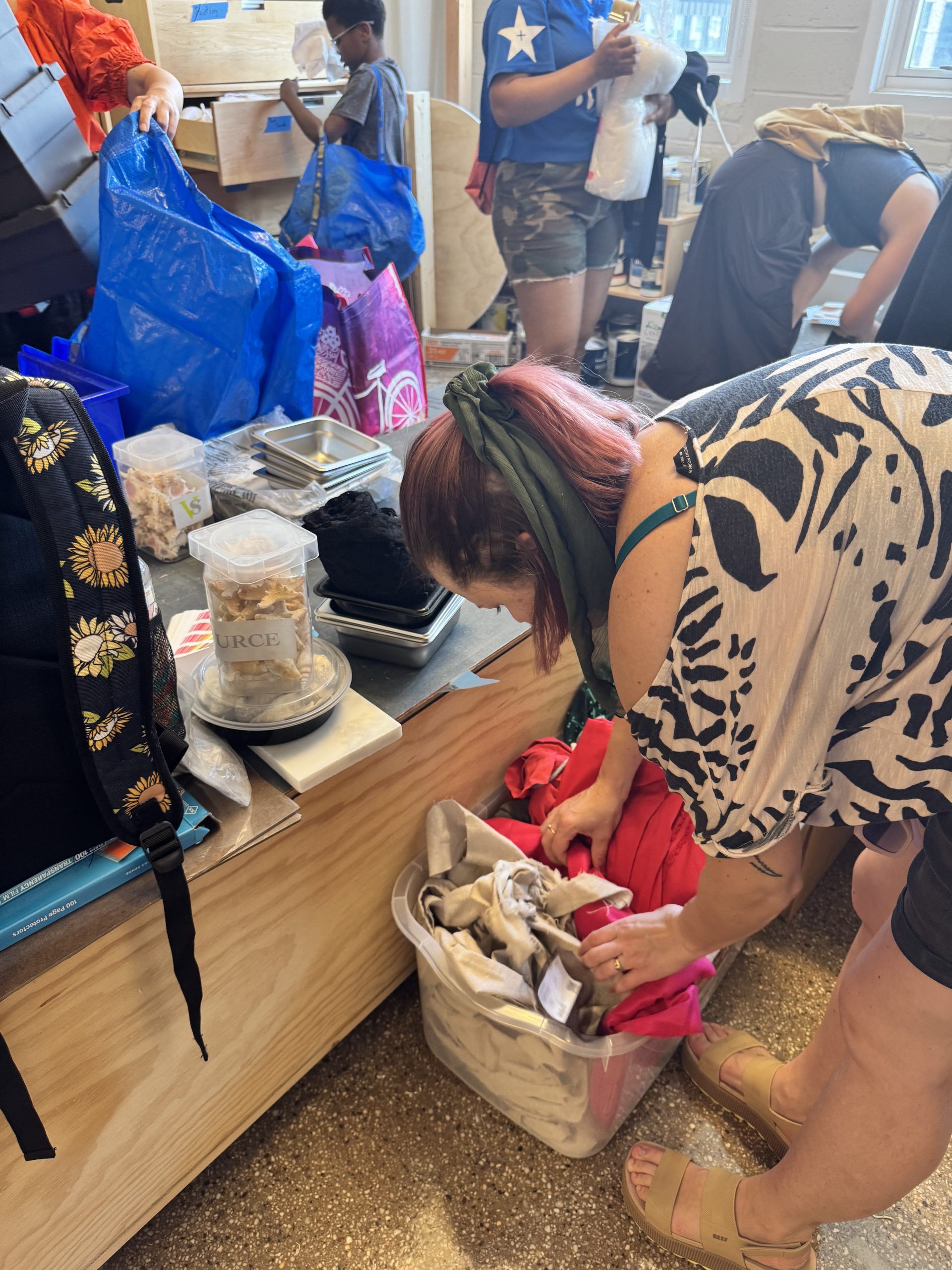 A woman with pink hair leans over to add to a bag filled with art materials as other artists look through cabinets of materials around her in the Art Pantry