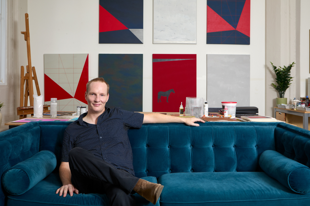 Artist Aaron Boucher seated on a blue velvet sofa in his studio, surrounded by geometric abstract paintings in red, blue, gray, and white, with art supplies and an easel nearby.