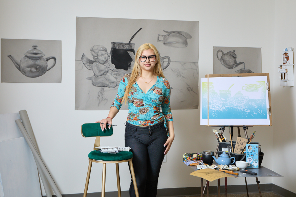 Artist Agustina Cueto stands in her ARTS 14C studio surrounded by large charcoal drawings of teapots and still-life objects, with drawing tools, ceramics, and a colorful work on an easel nearby.