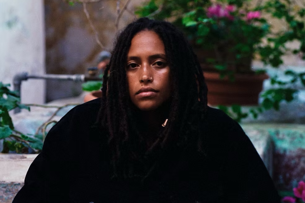 ARTS 14C Fellowship Artist Alba Roland Mejia looks directly at the camera in an outdoor portrait, framed by lush greenery and colorful flowers.