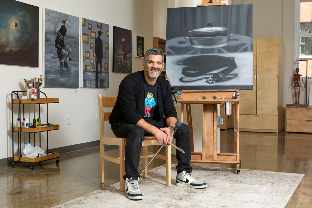 Artist Alex Martinez sits smiling in his ARTS 14C studio, holding paintbrushes beside a large monochromatic painting on an easel, with additional figurative paintings displayed across the walls behind him.