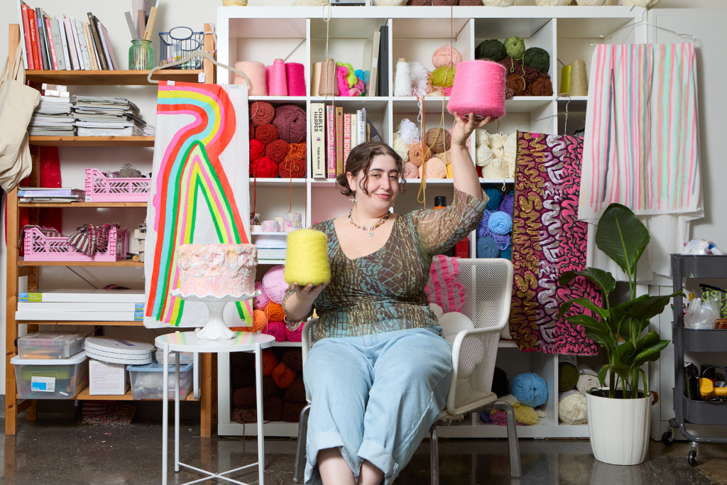 Visual artist Alexis Caruso seated in a colorful studio, playfully holding strands of yarn amid shelves filled with yarn, books, textiles, and vibrant fiber artworks.