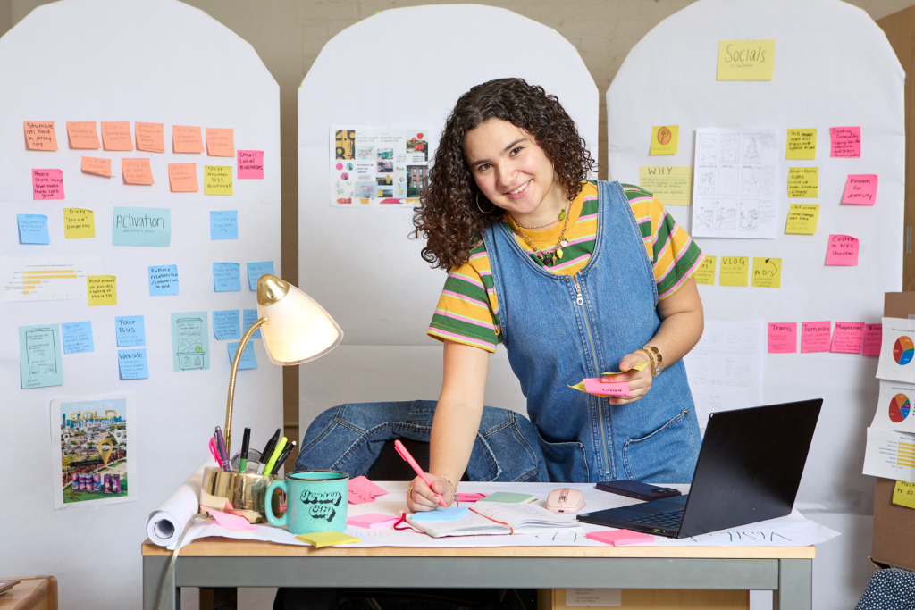 Special Project Artist Alyssa Dobles works at a desk covered with colorful sticky notes, notebooks, and a laptop, surrounded by large brainstorming boards filled with sketches, research, and ideas