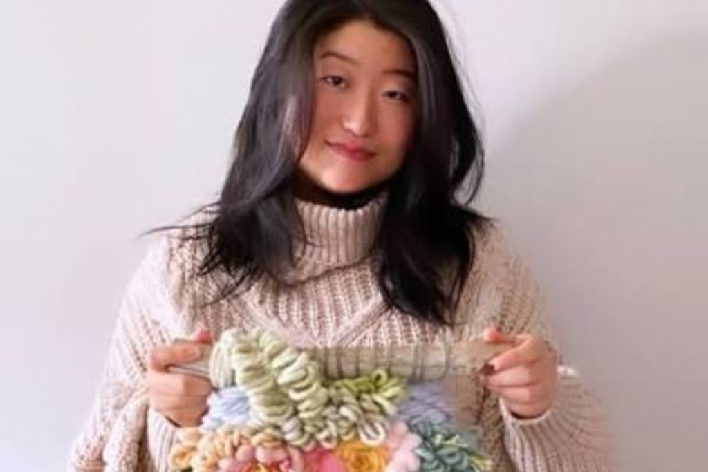 ARTS 14C Studio Artist Alyssa Ki holding a colorful textured fiber artwork while wearing a cream knit sweater against a light background.