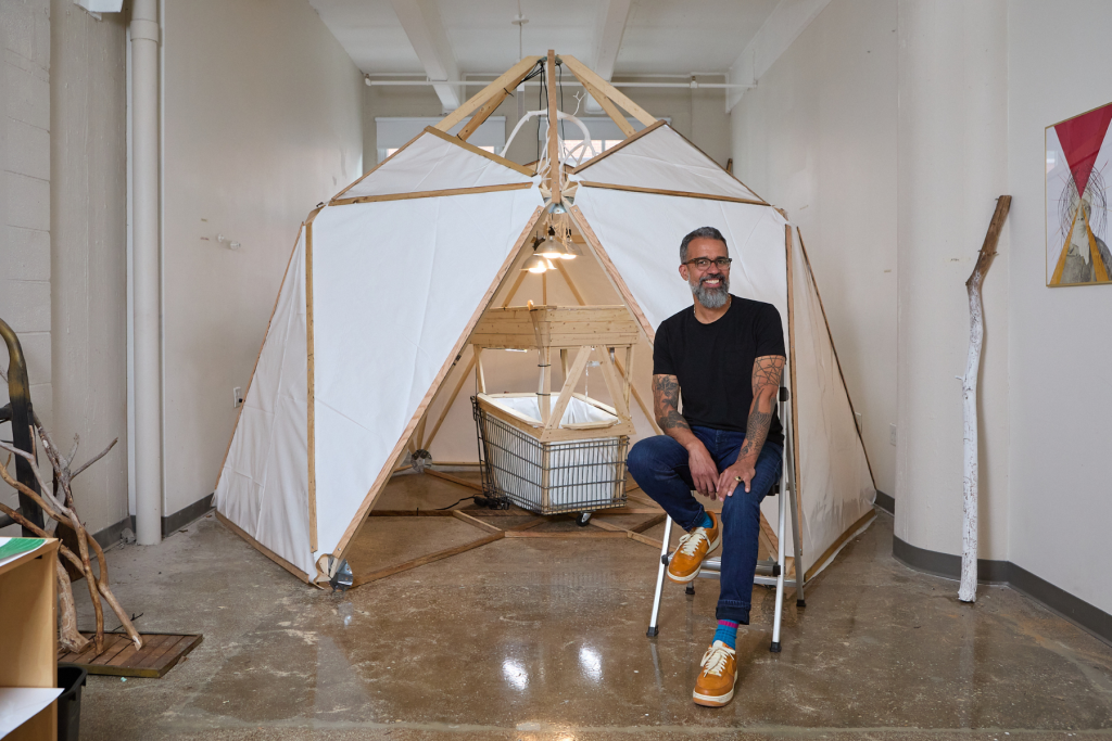 Artist Andrew Harrison sits smiling in his ARTS 14C studio beside a large, tent-like sculptural installation constructed from wood and white fabric.