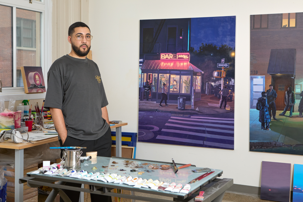 Artist Angel Duran stands in his ARTS 14C studio beside large paintings of nighttime urban scenes, with paint tubes, brushes, and a mixing palette spread across his worktable.