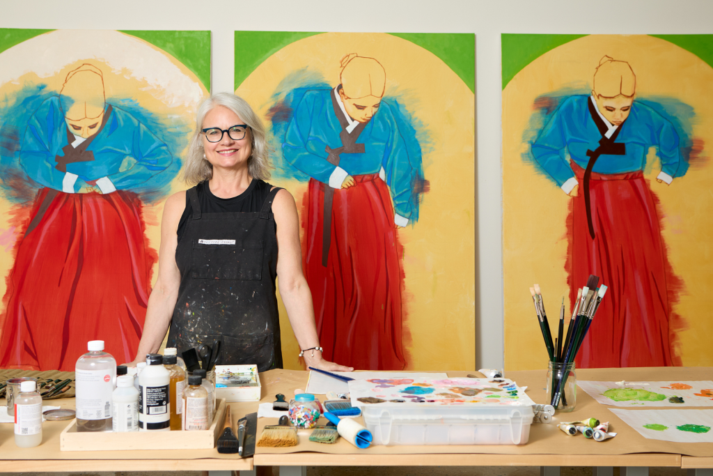 Artist Ann Antoshak smiles in her studio behind a worktable filled with paints, brushes, and art supplies, with three large paintings of figures in blue tops and red skirts displayed behind her.