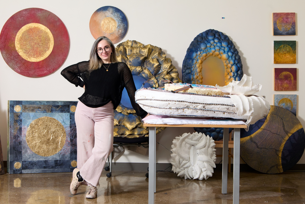 Artist Anna Collevecchio stands in her ARTS 14C studio surrounded by sculptural and mixed-media works in blue, gold, white, and red, many featuring circular forms and richly textured surfaces.
