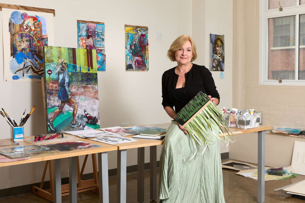Artist Anna Demovidova stands in her ARTS 14C studio holding a green mixed-media artwork, surrounded by colorful figurative paintings, works on paper, brushes, paints, and other materials from her creative practice.