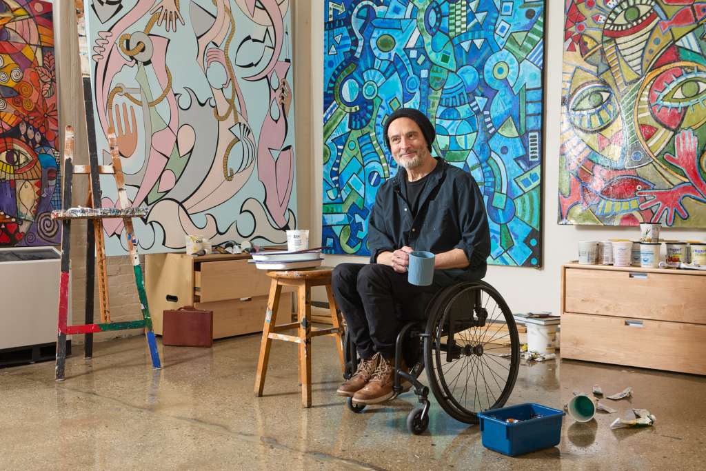 Artist Antonio Nogueira sits in a wheelchair holding a mug in his ARTS 14C studio, surrounded by large, vividly colored abstract paintings, an easel, paint supplies, and works in progress.