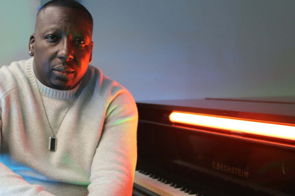 Portrait of a musician seated beside a grand piano, looking toward the camera as vivid red, green, and blue lighting creates a dramatic atmosphere across his face, white sweater, and the piano.