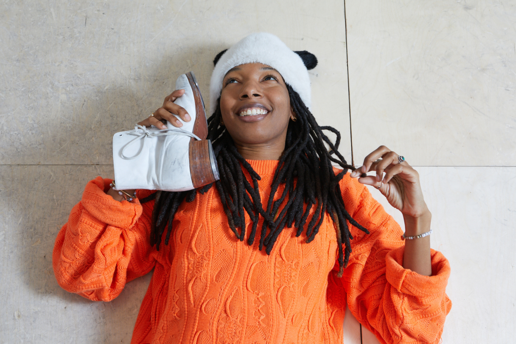 Artist Asha DaHomey in a portrait by Megan Maloy smiling in a bright orange sweater and fuzzy white hat, playfully holding a white boot to her ear while reclining against a light-colored wall.