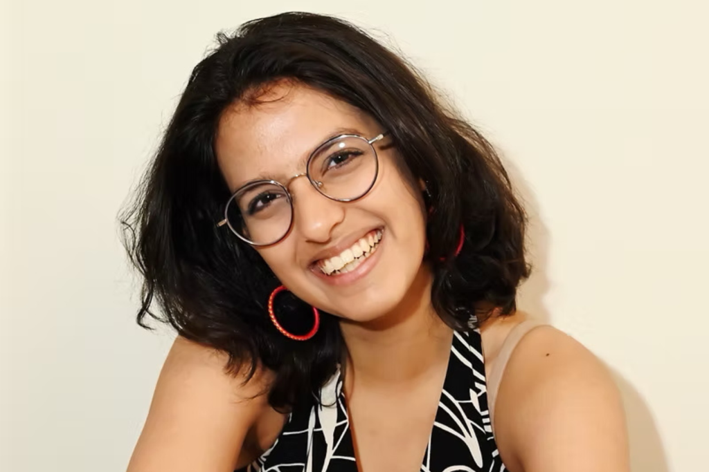 ARTS 14C Fellowship Artist Asta Trivedi smiles at the camera in a close-up portrait, wearing round glasses and red hoop earrings against a light background.