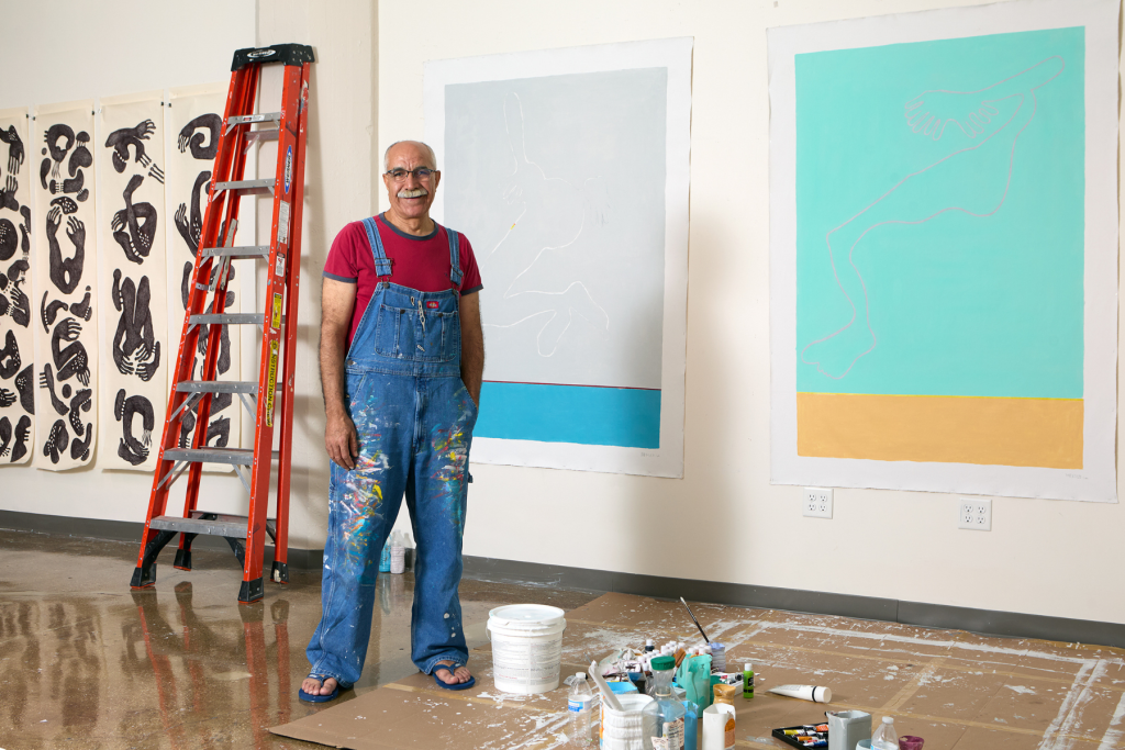Artist Ayman ElSamary stands in paint-splattered overalls in his ARTS 14C studio, surrounded by large figurative works, art supplies, and a red ladder.