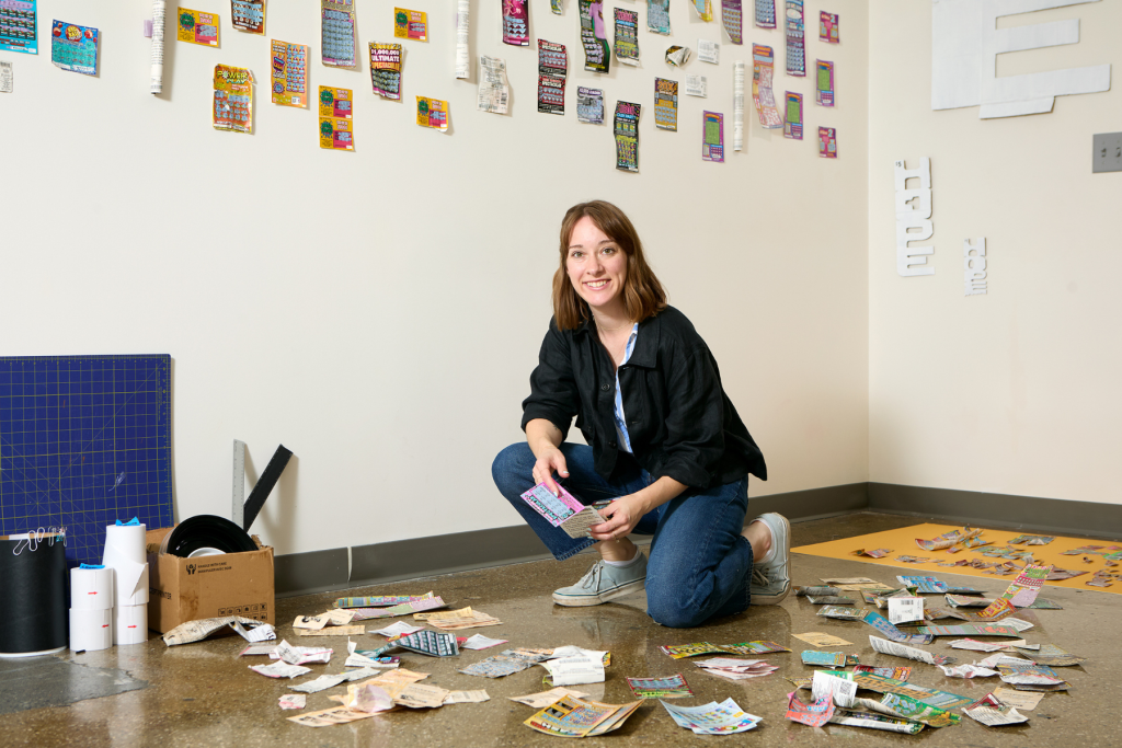 Artist Beth Fileti kneels in her ARTS 14C studio amid scattered lottery tickets and paper scraps, with rows of colorful tickets and sculptural paper forms displayed on the walls.