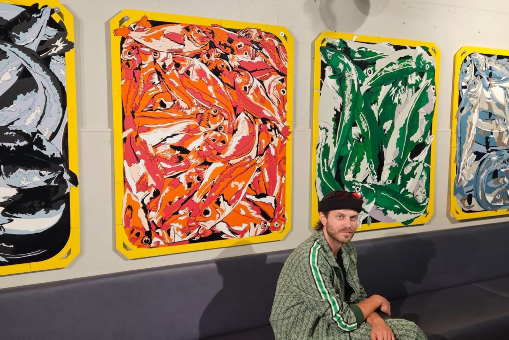 ARTS 14C Special Project artist Björgvin Jónsson seated beneath a series of vibrant, graphic artworks depicting densely layered fish in orange, green, blue, black, and white.