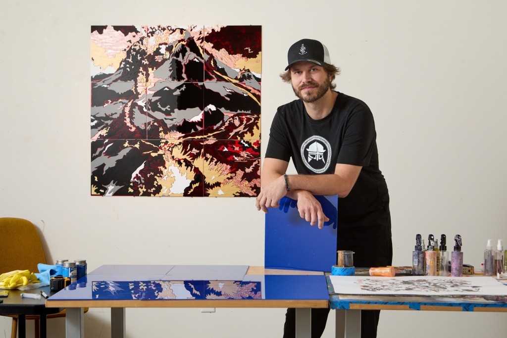 Artist Björgvin Jónsson stands in his ARTS 14C studio beside a graphic, multi-panel landscape artwork, with additional works in progress and art-making materials spread across nearby tables.