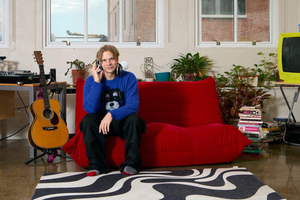 Musician Brandon Lowry (a.k.a. Sterling Fox, a.k.a. Baby Fuzz) sits on a bright red sofa in his ARTS 14C live/work studio, listening through headphones amid a guitar, DJ equipment, books, plants, and a vintage television.