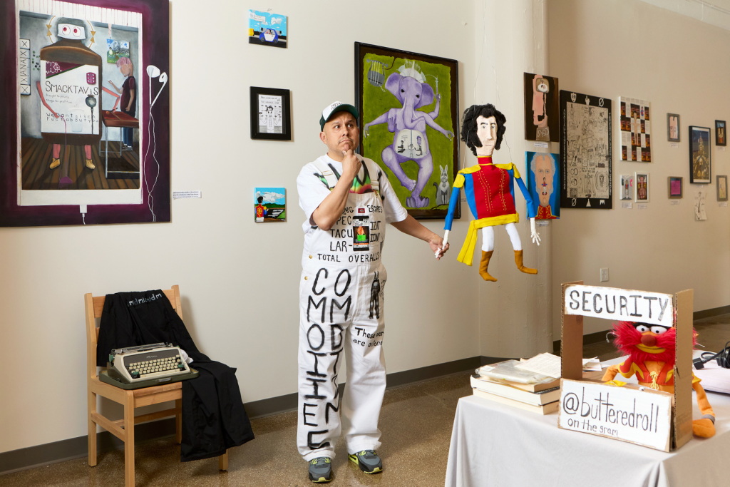 Artist Buttered Roll stands in a gallery surrounded by playful mixed-media artworks, holding a colorful handmade puppet beside a table featuring a puppet “security” booth.