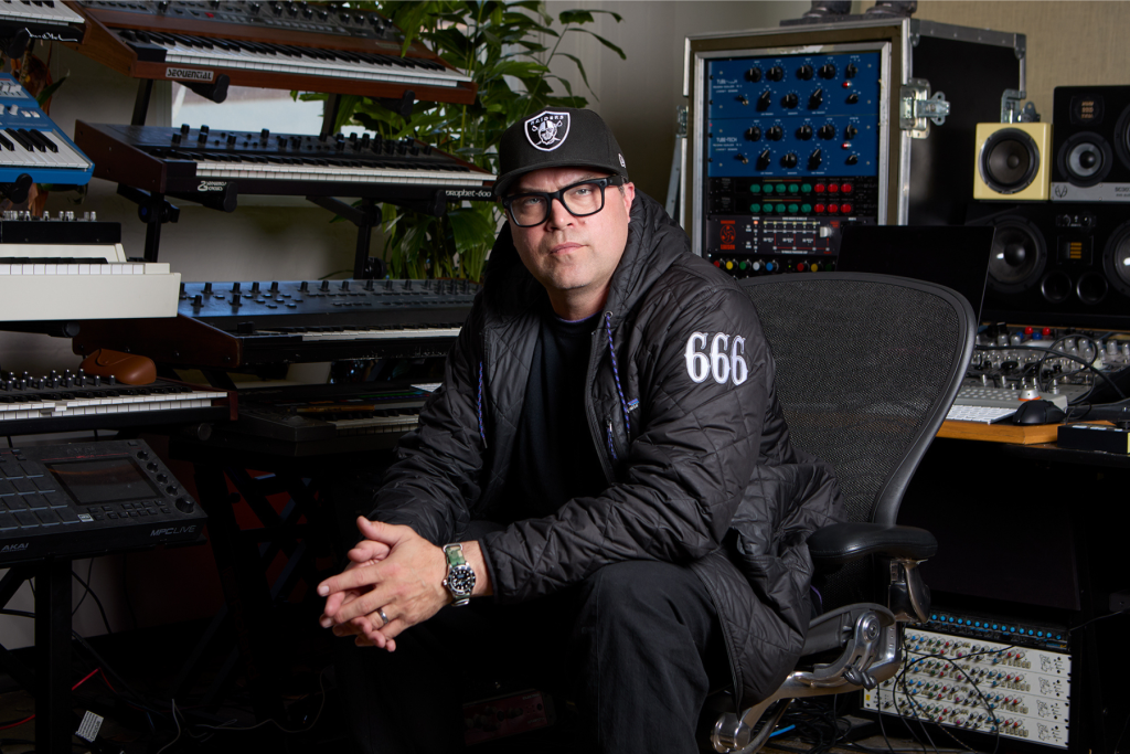 Chris Sieg sits in his music production studio at ARTS 14C surrounded by synthesizers, keyboards, speakers, and audio equipment, wearing a black cap, glasses, and jacket.