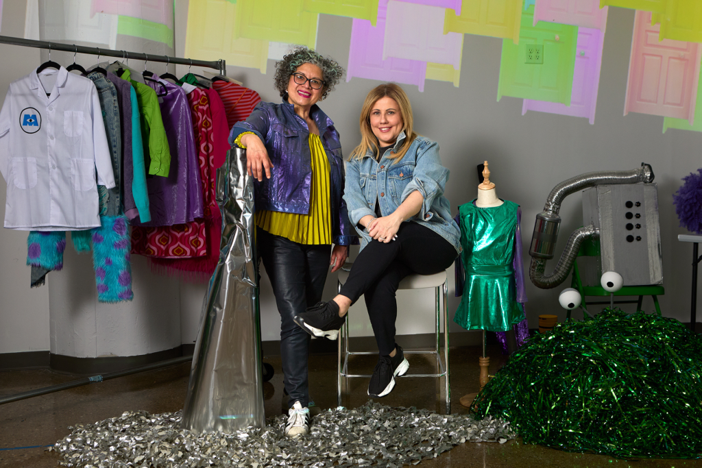 Melida Rodas and Nicole Oliver from City Kidz Playhouse pose together in a colorful studio surrounded by costumes, metallic fabrics, sculptural props, and a dress form, with brightly colored door shapes projected on the wall behind them.