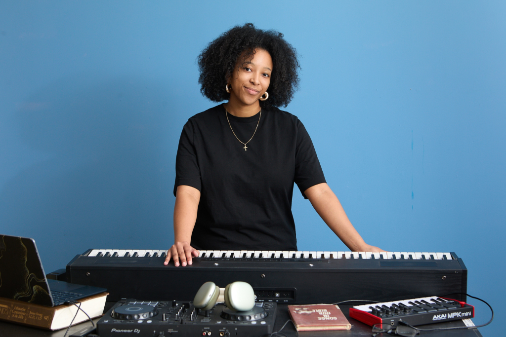 Composer Danae Venson stands behind a keyboard and DJ equipment against a blue wall, surrounded by tools of her creative practice, including a laptop, headphones, mixer, MIDI controller, and songbook.