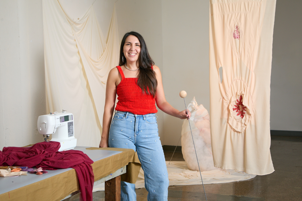 Artist Diana Jean Puglisi stands smiling in her ARTS 14C studio, surrounded by suspended textile works and fabric, with a sewing machine on her worktable.