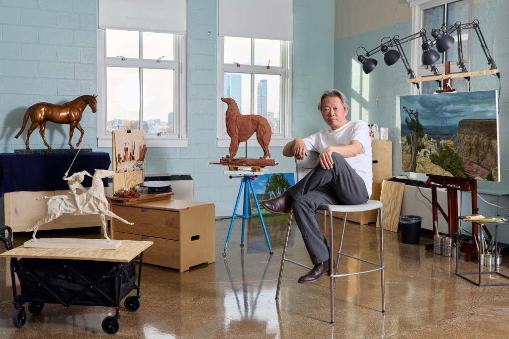Artist Dong Yang seated in a bright studio surrounded by horse sculptures, a clay horse in progress, a landscape painting, and art-making tools.