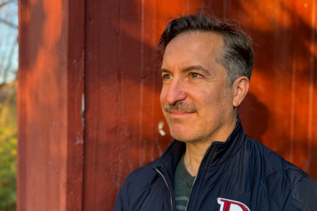 ARTS 14C Fellowship Artist Drew Pisarra in an outdoor portrait, wearing a dark quilted jacket and looking off-camera against a warmly lit red wooden background.