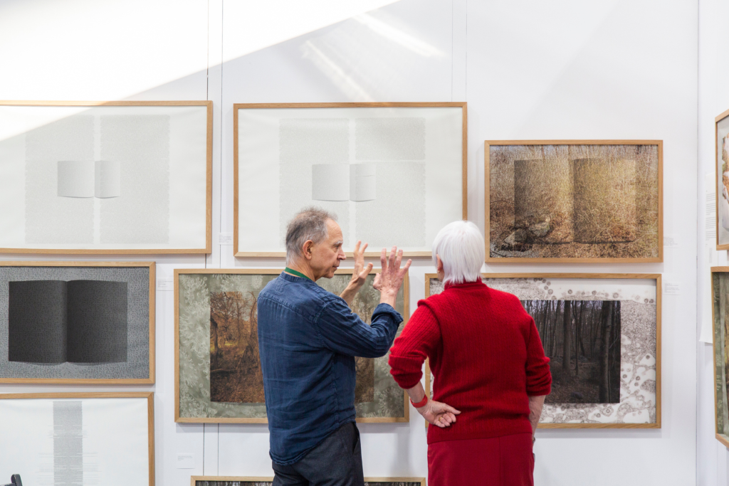 Artist Edward Fausty gesturing while discussing framed mixed-media artworks with a visitor in a bright gallery exhibition.
