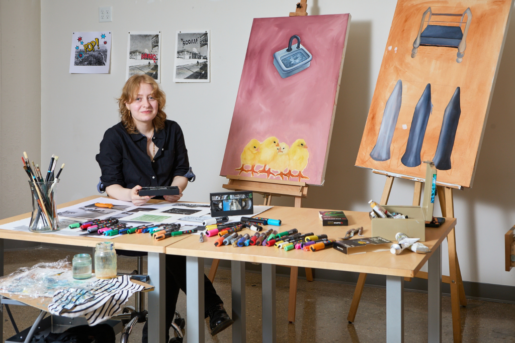 Artist Elaf Hussein sits at a worktable in her ARTS 14C studio, surrounded by colorful markers, brushes, paintings on easels, and graphic works displayed on the wall.