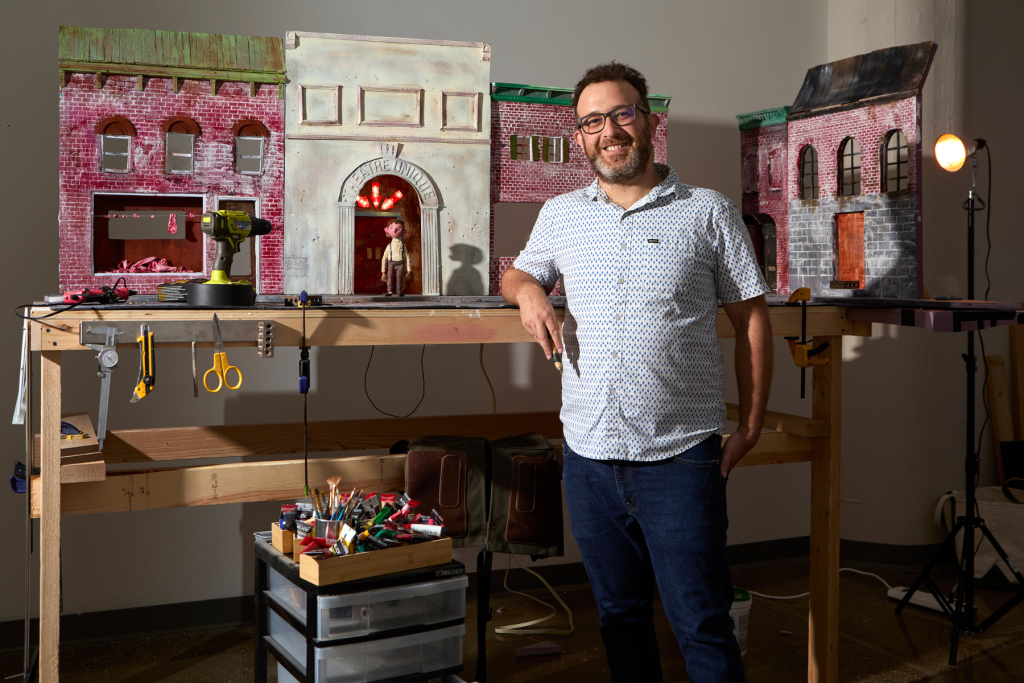 Stop motion filmmaker Elliot Lobell stands in his ARTS 14C studio beside a detailed miniature theater set, surrounded by model-making tools, paints, lighting equipment, and handcrafted architectural elements.