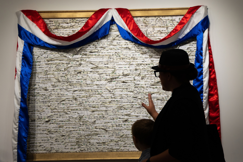 Artist Ellis Angel is seen in shadow in front of her large mixed-media artwork made from shredded U.S. currency and the shredded tax returns of Donald Trump, framed by draped red, white, and blue fabric in a gallery.