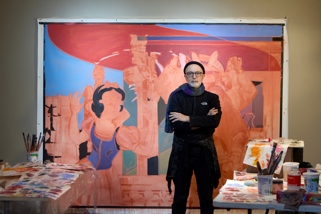 ARTS 14C Studio Artist Farsad Labbauf standing with arms crossed in his studio before a large-scale painting in coral, blue, and muted tones, surrounded by paint-covered worktables, brushes, and art supplies.