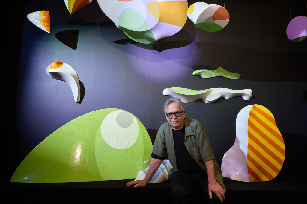 Project 14C artist George Goodridge seated among colorful abstract sculptural forms in green, orange, yellow, purple, and white against a dark gallery backdrop.