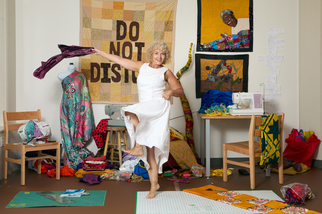 ARTS 14C Special Project artist HAMEWS posing playfully in her textile studio, surrounded by colorful fabrics, sewing machines, dress forms, quilting tools, and vibrant fiber artworks.