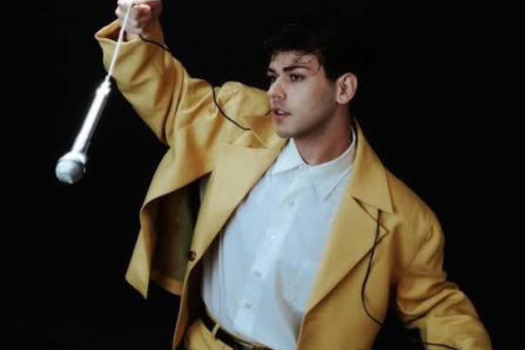 ARTS 14C 12-month Fellowship artist Haven Bell holding a suspended microphone while wearing a mustard-yellow suit against a black background.