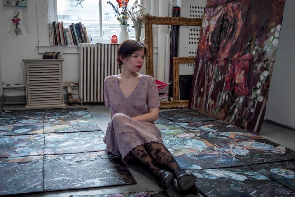 ARTS 14C Fellowship Artist Helen Wordsworth seated on the floor of her studio, surrounded by large, richly layered paintings arranged across the floor and leaning against the wall.