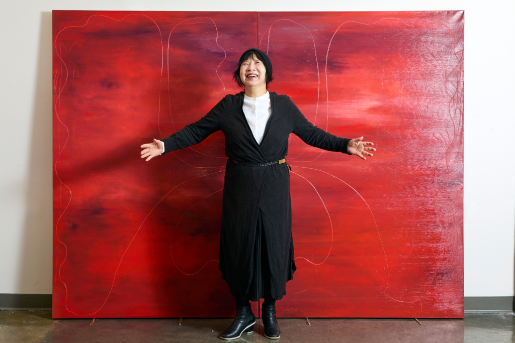 Artist Heryun Kim stands with arms outstretched in her ARTS 14C studio, smiling in front of a monumental two-panel abstract painting in deep, vivid shades of red.