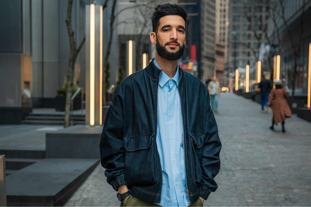 ARTS 14C Special Project artist Jani Grigoryan standing on a city plaza, wearing a dark jacket and light blue shirt, with illuminated columns and buildings in the background.
