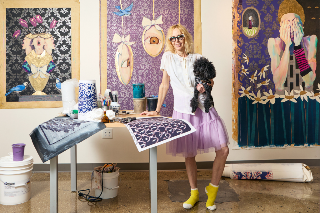 ARTS 14C Fellowship artist Flying Knight standing in her studio holding a small black dog, surrounded by large, richly patterned figurative artworks in purple, blue, gold, and black.