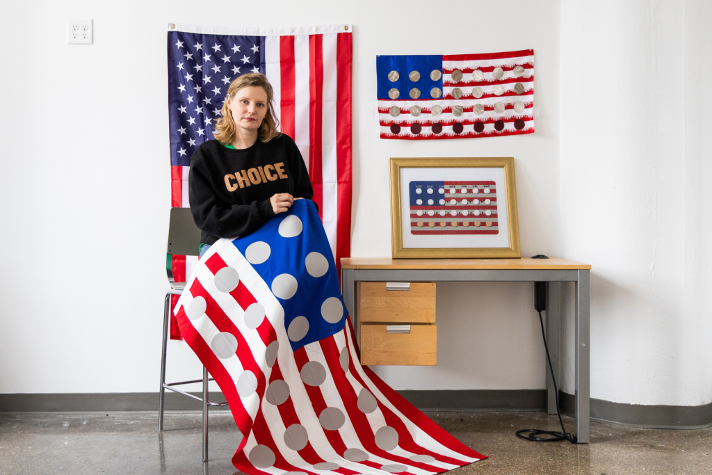 Artist Katrina Majkut sits in her ARTS 14C studio surrounded by works that reinterpret the American flag, holding a red, white, and blue textile piece covered with large circular forms.