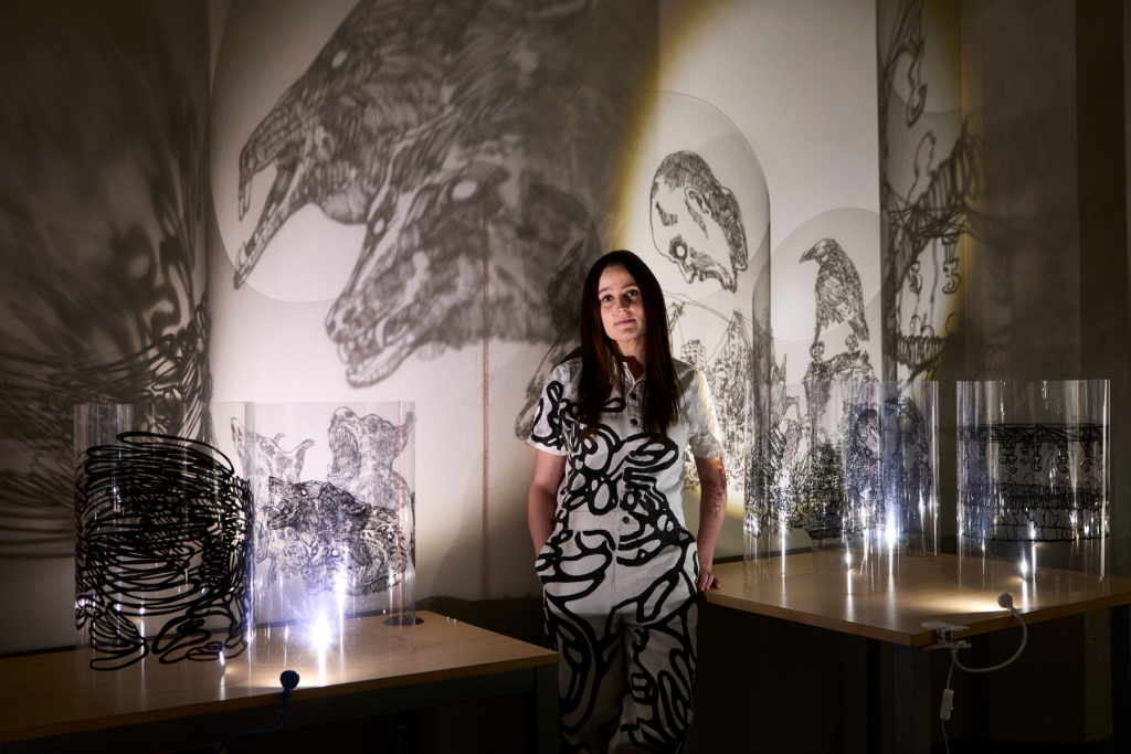 Artist Katya Muromtseva stands in her darkened ARTS 14C studio among illuminated transparent sculptures whose intricate black drawings cast dramatic projections of animals, figures, and architectural forms across the walls.