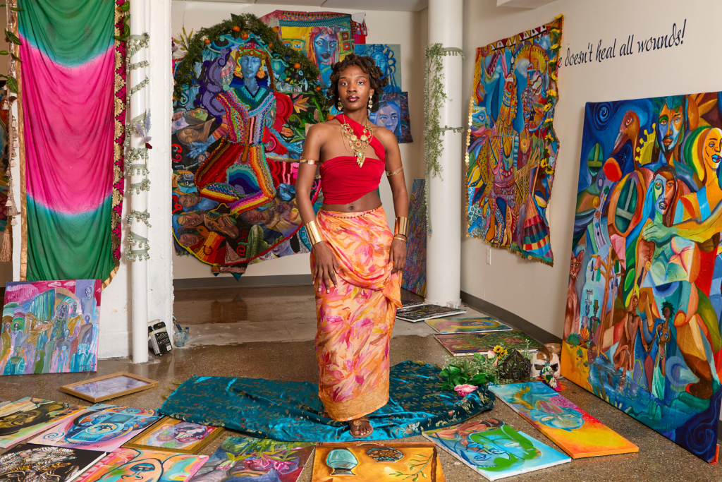 Artist Kayla (Eleven) Muldrow stands in her ARTS 14C studio surrounded by vivid, large-scale paintings and textile works filled with expressive figures, bold patterns, and saturated color.