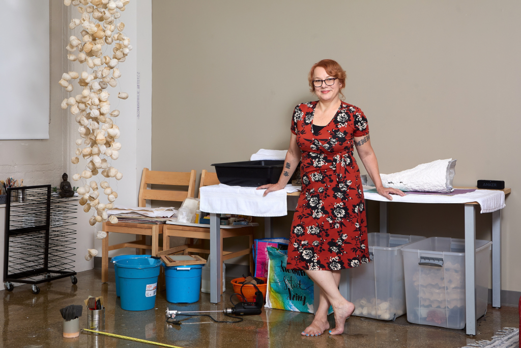 Artist Keely McCool stands barefoot in her ARTS 14C studio beside a worktable with sculptural materials, tools, and containers, while a cascading white organic sculpture hangs nearby.