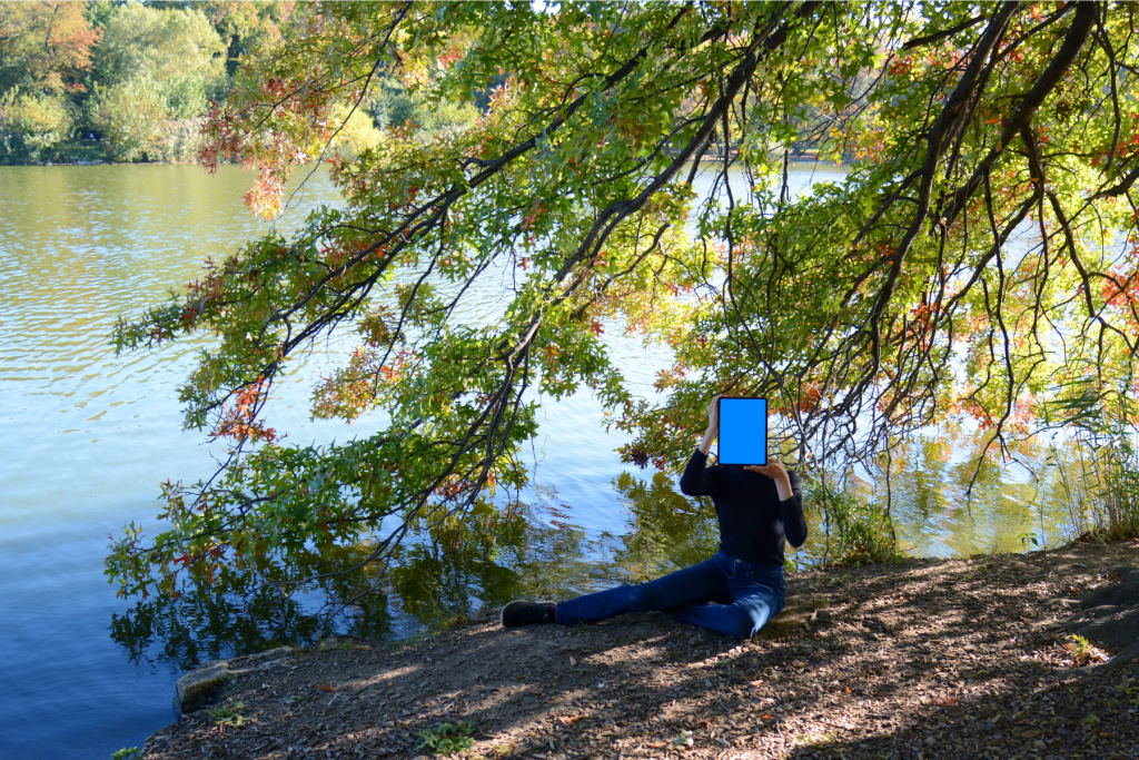 ARTS 14C Special Project artist LIU ENTUNG seated beneath leafy tree branches beside a lake, holding a tablet with a vivid blue screen in front of her face.