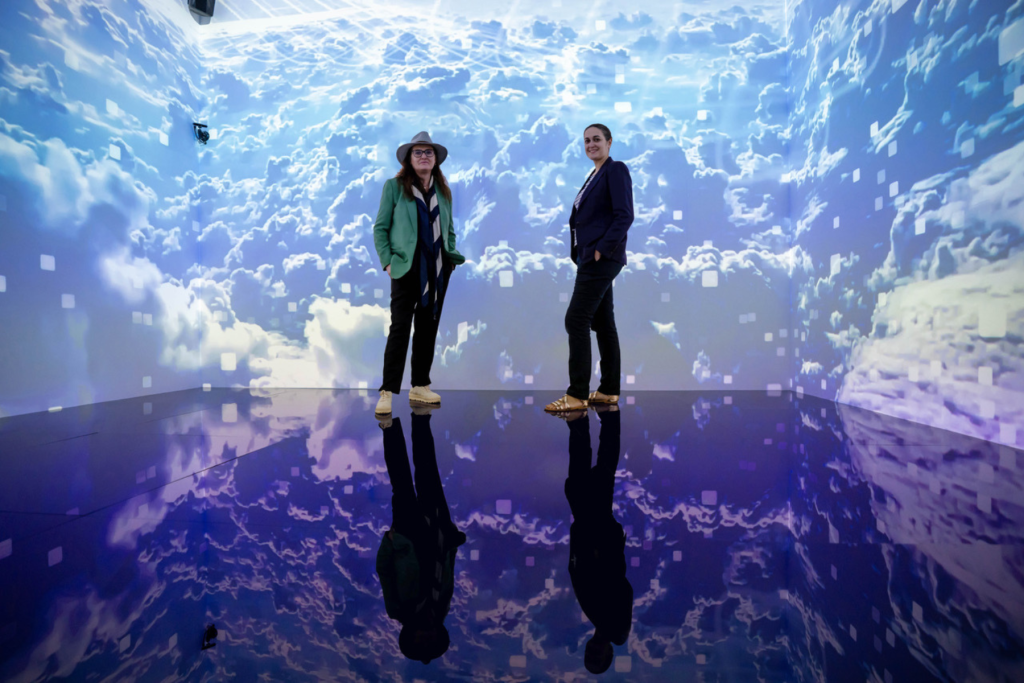 Artists Laia Cabrera and Isabelle Duverger stand within an immersive installation of luminous blue skies and clouds projected across the walls and reflected on a glossy floor.